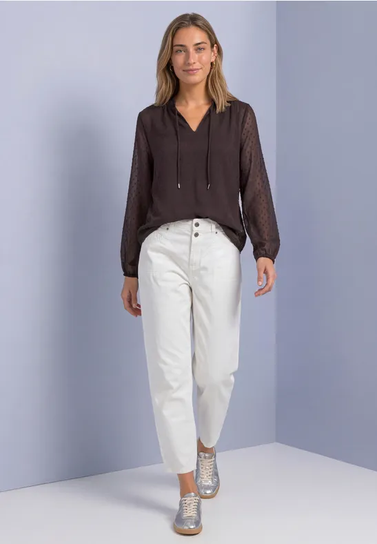 Femme en chemisier sombre, pantalon blanc taille haute et chaussures argentées, posant devant un mur lilas.