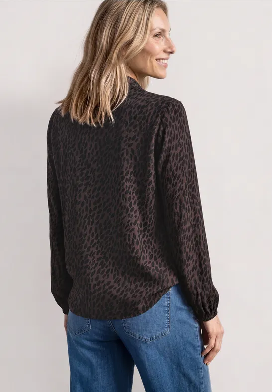 Frau von hinten in dunkelbrauner Leoprint-Bluse und blauen Jeans vor hellgrauem Hintergrund.
