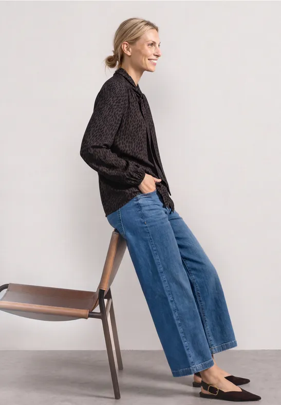 Seitlich abgebildete Person mit schwarzem Hemd, weitem Jeans, schwarzen Loafers lehnt an einem Holzstuhl.