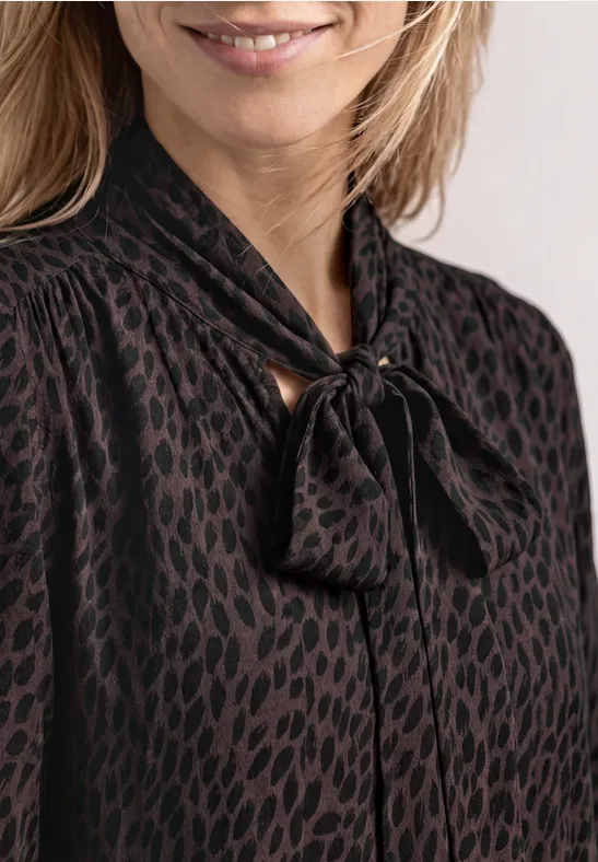 Frau in schwarzer Bluse mit dezentem Leopardenmuster und Schleifenkragen.