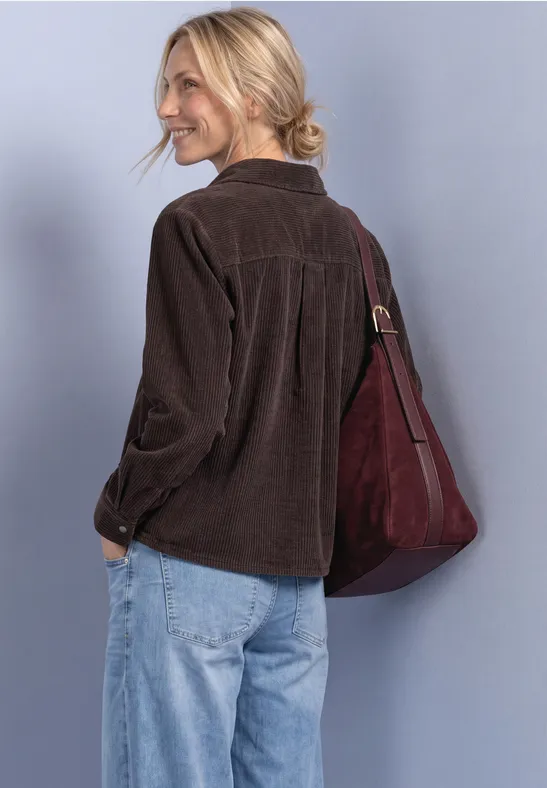 Eine Frau in brauner Cordjacke, blauen Jeans und roter Tasche, lächelnd, von hinten aufgenommen.