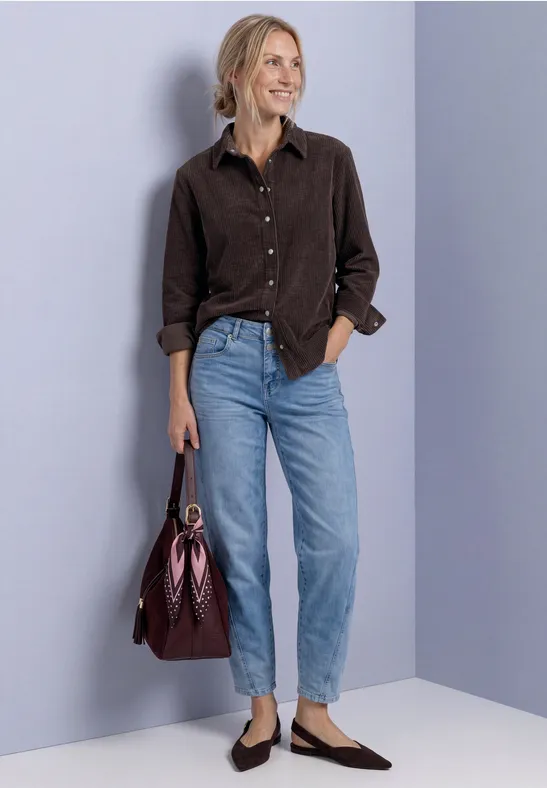 Frau in braunem Hemd und blauen Jeans mit burgunderfarbener Tasche lächelnd vor grauem Hintergrund.