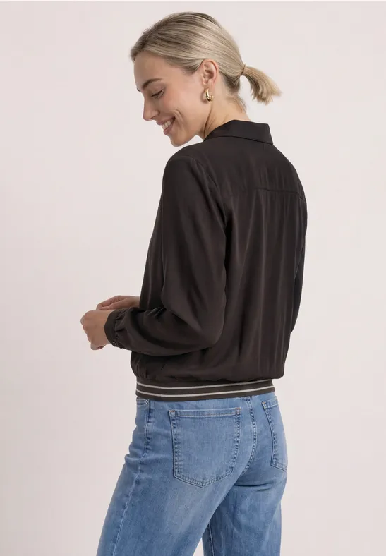 Frau von hinten in schwarzer Bomberjacke und blauen Jeans, lächelnd, Haare im Pferdeschwanz.