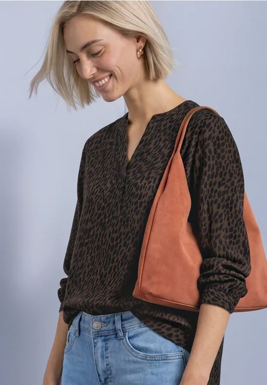 Eine Frau in gemustertem Pullover, Jeans und braun-orangener Tasche lächelt und blickt nach unten.