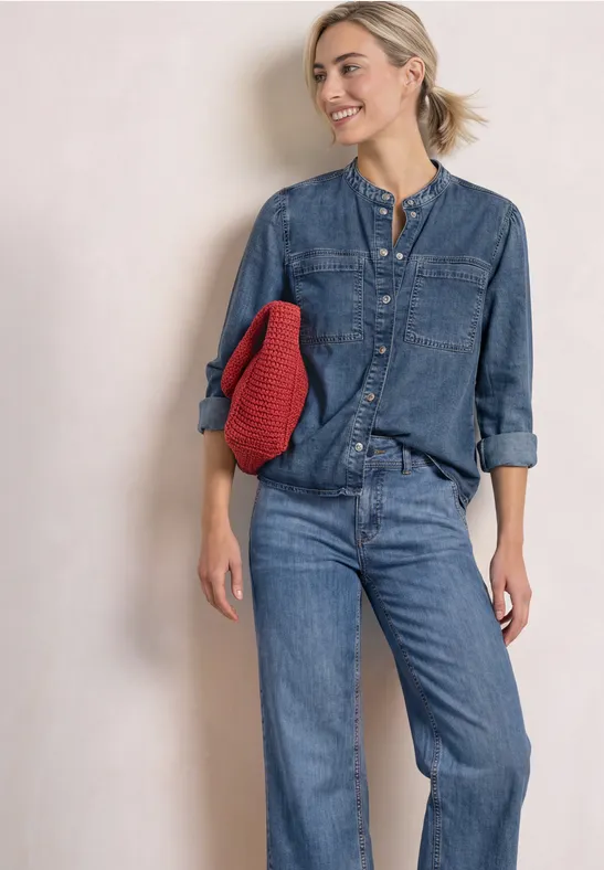 Eine Frau in blauem Jeans-Outfit lehnt lächelnd an einer Wand und hält einen roten Pullover.