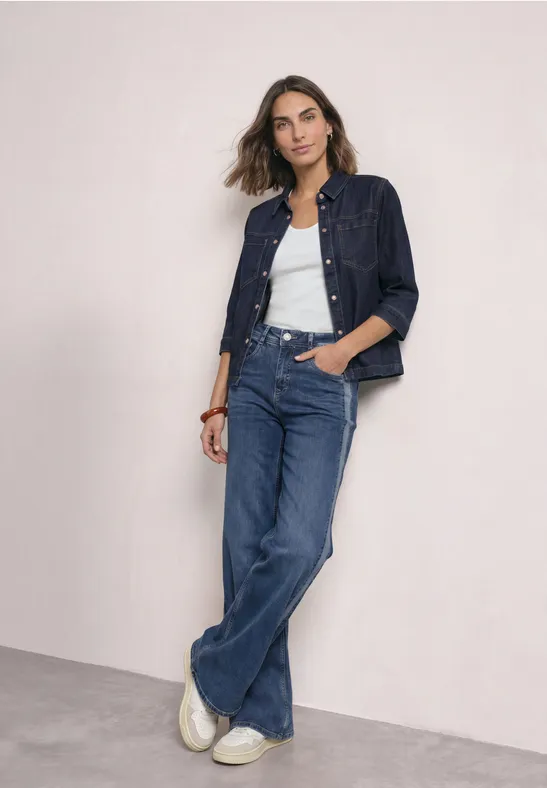 Frau in Jeansjacke, weißem Top und weit geschnittenen Jeans lehnt lässig an einer hellen Wand.