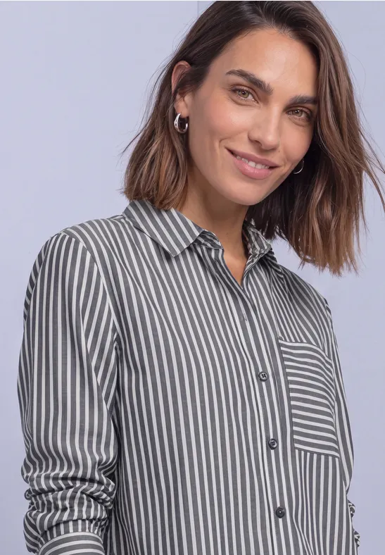 Une femme souriante porte une chemise rayée à manches longues avec poche poitrine, fond clair.