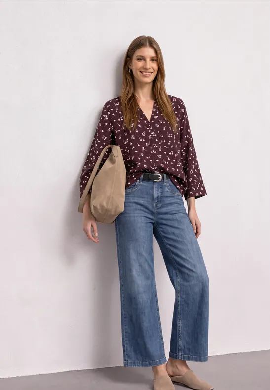 Eine lächelnde Frau trägt eine weinrote Blumenbluse, weite Jeans und eine große beige Tasche über der Schulter.