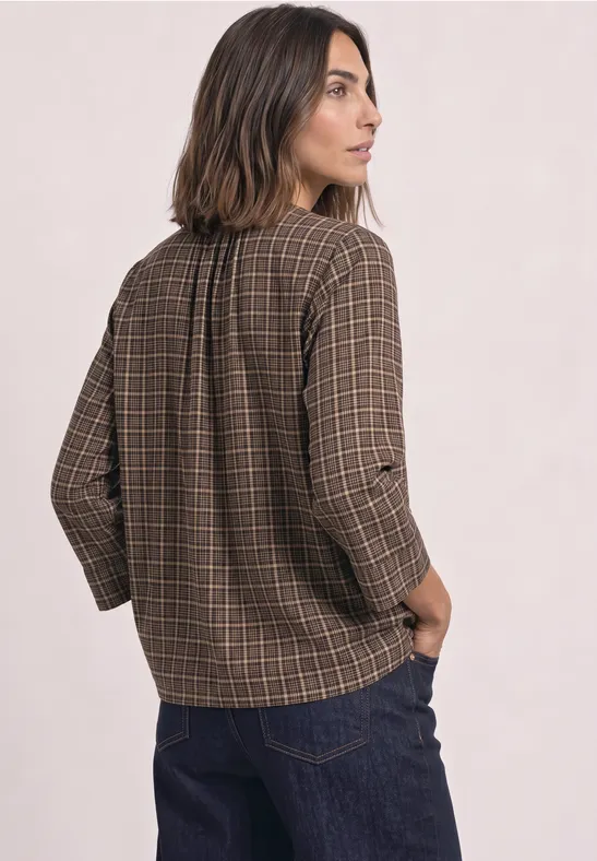 Eine Frau steht im Profil, trägt karierte braune Bluse mit langen Ärmeln und blaue Jeans.