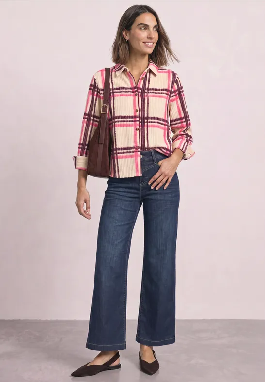 Frau in karierter Bluse, weiten Jeans und schwarzen Ballerinas trägt eine braune Schultertasche und steht lässig.