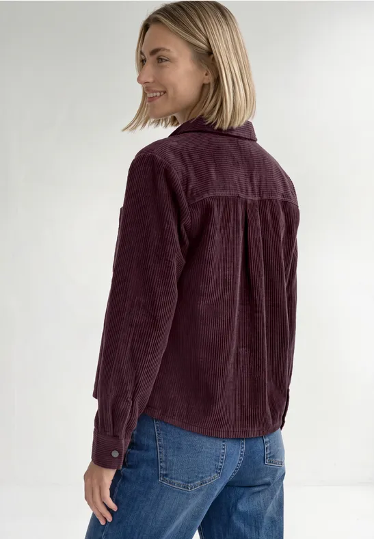 Frau mit kurzem blondem Haar trägt auberginefarbenes Cordhemd und blaue Jeans, lächelt seitlich zur Kamera.