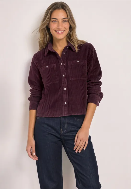 Lächelnde Frau in violetter Cordjacke und dunklen Jeans.
