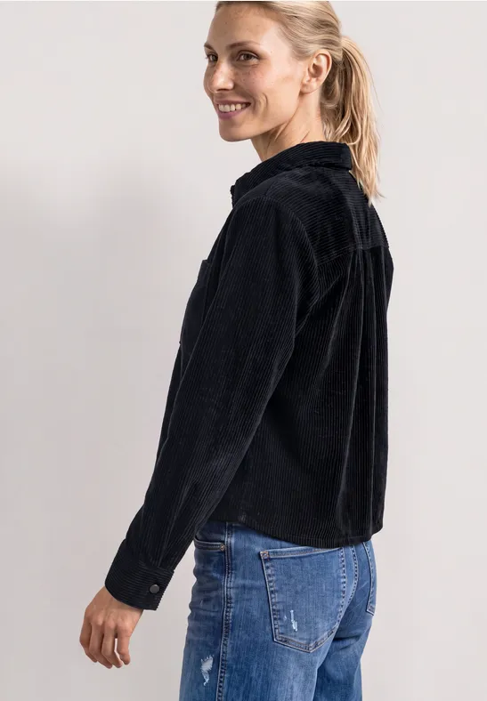 Eine Frau im schwarzen Cord-Jacket und blauer Jeans lächelt seitlich in die Kamera.