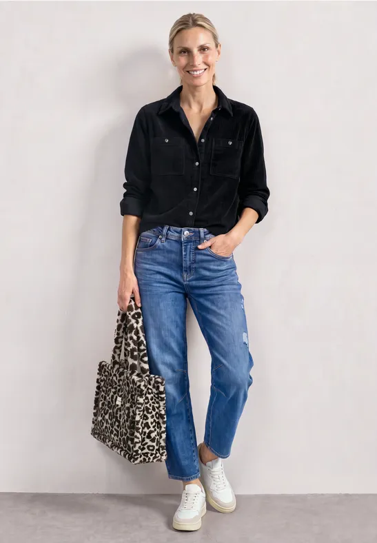 Eine Frau in schwarzem Hemd, blauen Jeans und weißen Sneakern, hält eine Leo-Tasche, lehnt an weißer Wand.