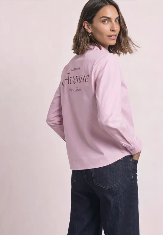 Eine Frau trägt eine rosa Jacke mit Schriftzug und blaue Jeans, schaut über die Schulter.