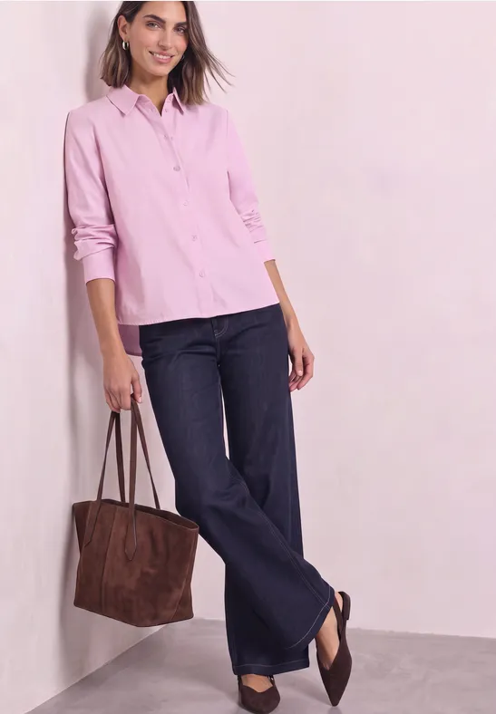 Frau trägt rosa Hemd, weite Jeans und braune Tasche, steht lässig an einer Wand.