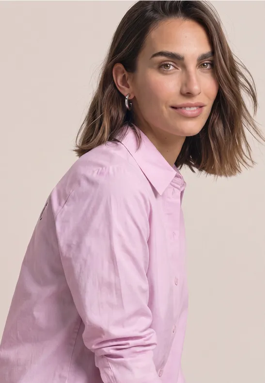 Eine Frau in rosa Hemd sitzt seitlich, blickt freundlich in die Kamera.