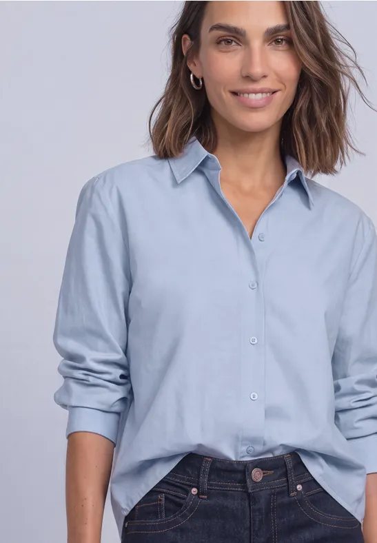 Vrouw in oversized lichtblauw hemd, mouwen opgestroopt, half ingestopt, met een zachte glimlach.