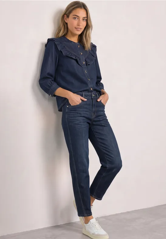 Eine Frau lehnt an einer Wand, trägt ein dunkles Denim-Outfit aus Hemd und Hose und schaut lässig in die Kamera.