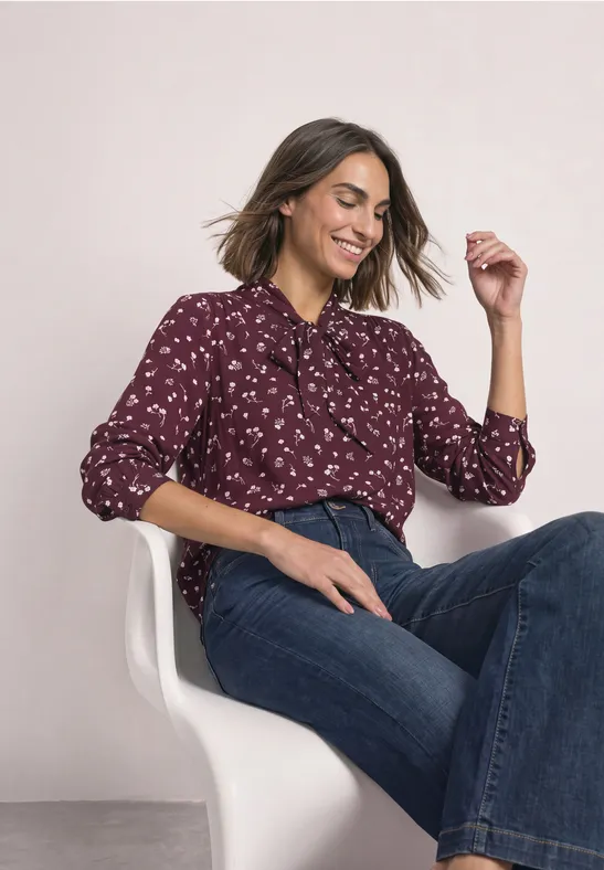 Frau in bordeauxroter Bluse mit Blumenprint und weiten Jeans sitzt lächelnd auf einem weißen Stuhl.