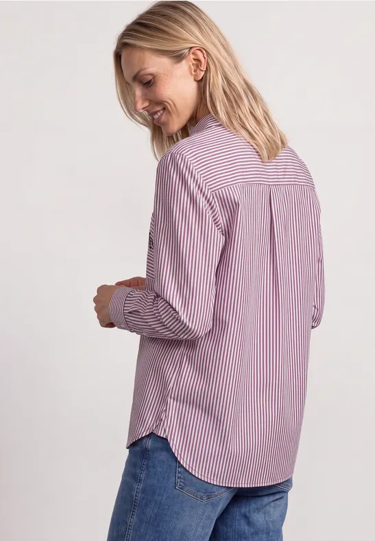 Une femme blonde porte une chemise rose et blanche à rayures et un jean, vue de profil, elle sourit.