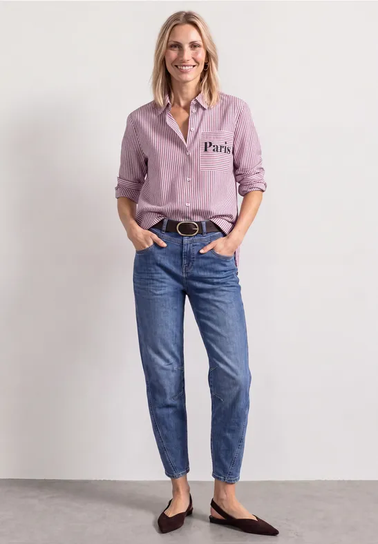 Femme en chemise rose, jean bleu et ballerines noires devant un fond neutre.