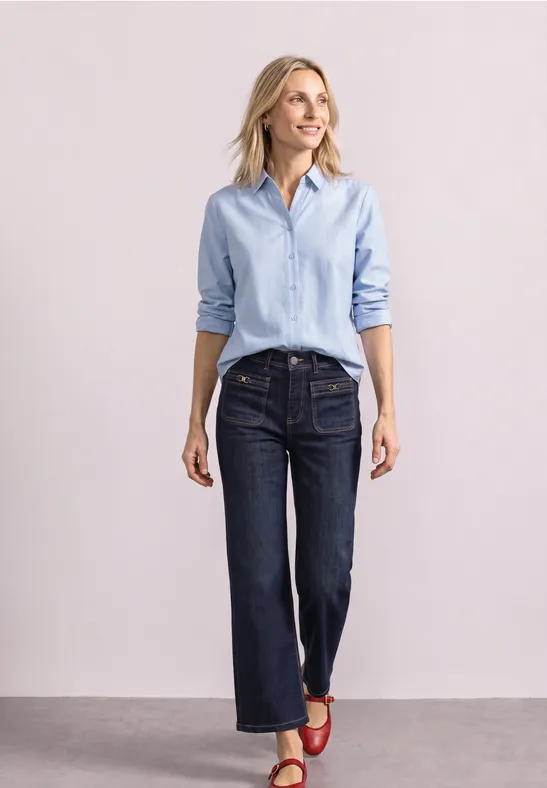 Vrouw in lichtblauw blouse, donkere jeans en rode sandalen voor een neutrale achtergrond.
