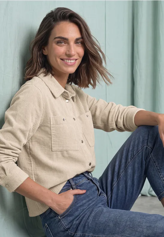 Een vrouw met bruin haar zit lachend voor een lichte achtergrond in een beige blouse en jeans.