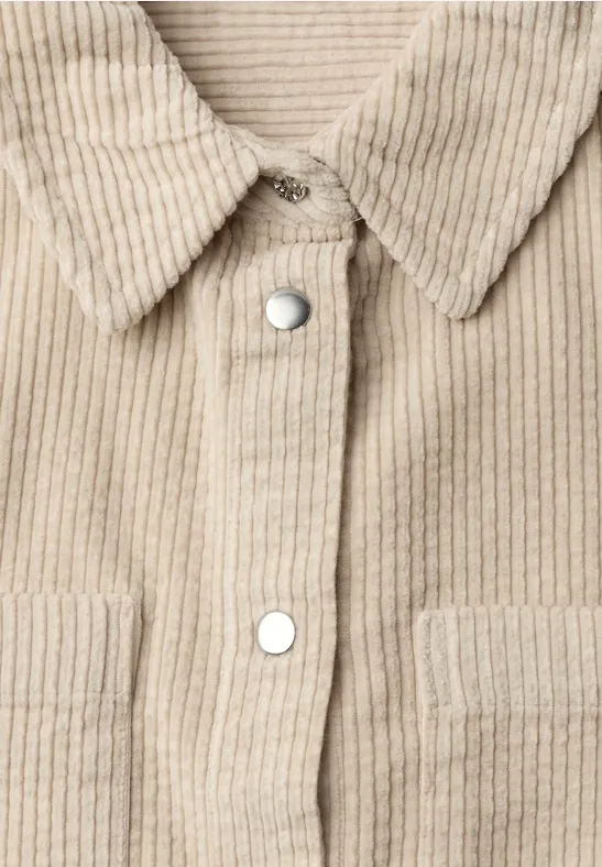 Close-up van voorkant van beige corduroy-overhemdjas met zilverkleurige drukknopen en opgezette zakken.