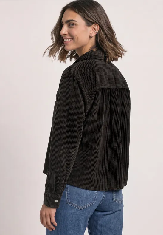 Eine lächelnde Frau in seitlicher Ansicht trägt eine braune Cordjacke und blaue Jeans.