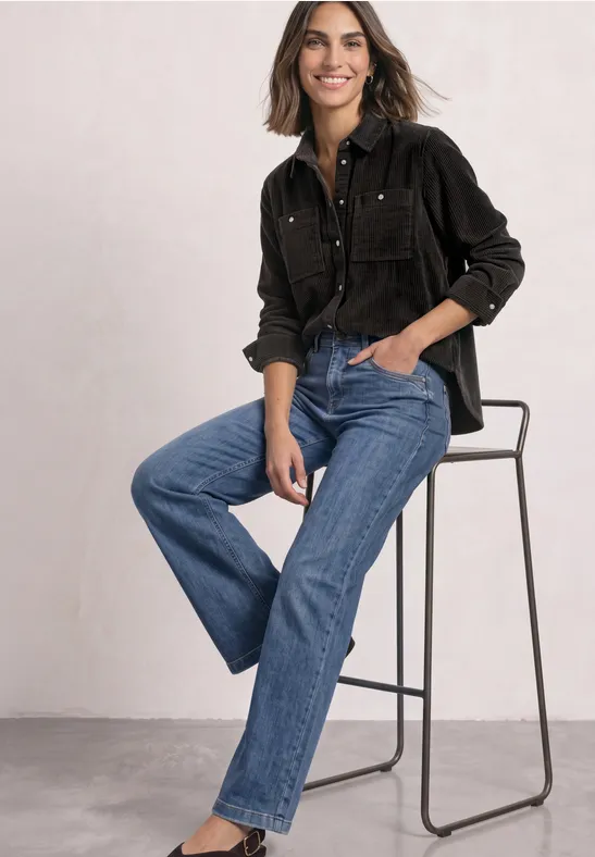 Frau auf hohem Stuhl in schwarzer Bluse und blauen Jeans, sie sitzt entspannt und lächelt.