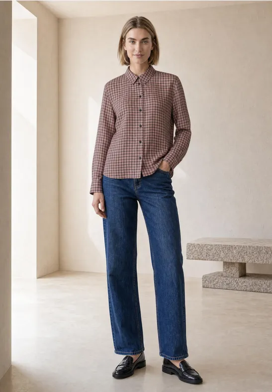 Eine Frau steht in minimalistischem Raum, trägt karierte Bluse, gerade geschnittene Jeans und schwarze Loafers.
