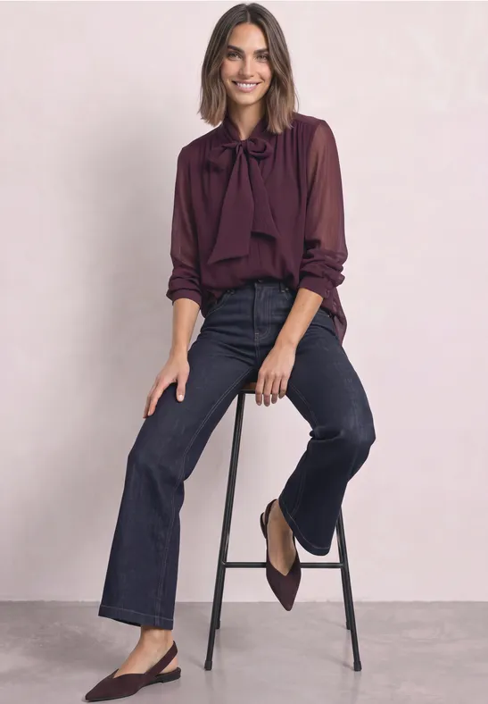 Dame sitzt auf Hocker, trägt weinrote Bluse mit Schleife, dunkelblaue Jeans und Slingback-Pumps.