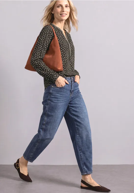 Frau in schwarzer gepunkteter Bluse, blauen Jeans, schwarzen Ballerinas und brauner Schultertasche vor einfarbiger Wand.