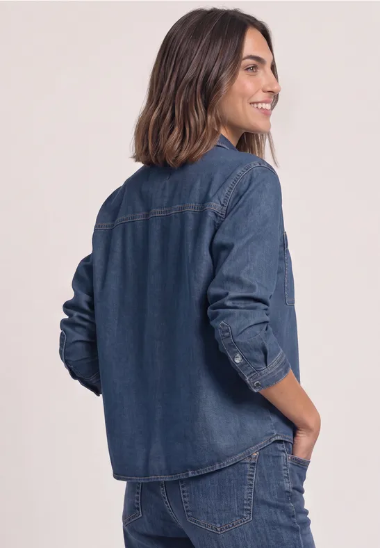 Eine Frau im dunkelblauen Denim-Set (Hemd & Hose), die seitlich lächelnd mit Hand in der Hosentasche steht.