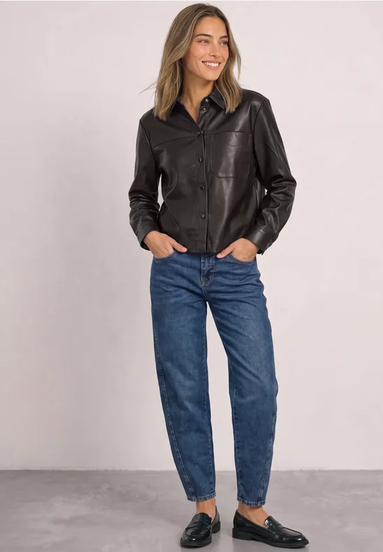 Frau in schwarzer Lederjacke, blauen Jeans und schwarzen Loafers, lächelnd mit Händen in den Taschen.