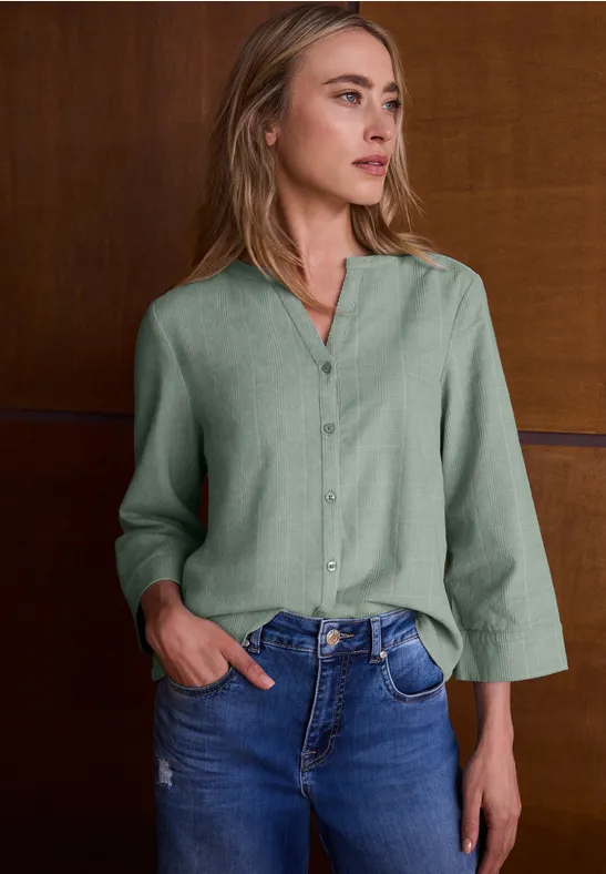Frau mit grüner Bluse und Jeans lehnt an einer Holzwand, Blick nach rechts.