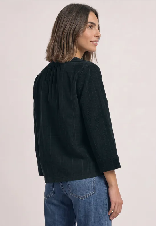 Frau steht seitlich abgewandt, trägt schwarze Bluse mit Struktur und blaue Jeans, schaut über die Schulter.