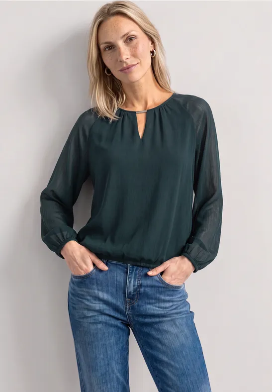 Vrouw in groene blouse en spijkerbroek tegen neutrale achtergrond, handen in de zakken.