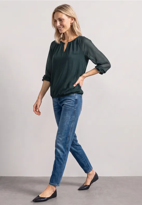 Vrouw in groene blouse en blauwe jeans, glimlachend poseert ze voor een witte achtergrond.