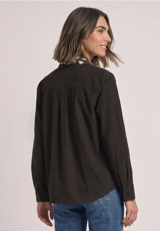 Eine Frau trägt eine dunkelbraune Cord-Hemdjacke und Jeans, sie blickt seitlich über die Schulter.