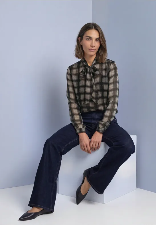 Vrouw in geruit bloes met strik, donkere flared jeans en zwarte pumps, zittend op een grijs blok.