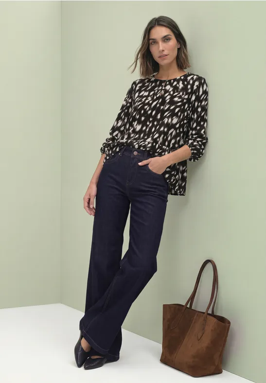 Eine Frau trägt eine schwarz-weiß gemusterte Bluse, dunkelblaue Hosen und eine braune Tasche.