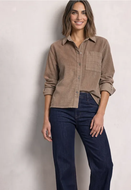 Frau in lässigem braunen Cord-Hemd und dunklen Jeans, stehend vor heller Wand, lächelnd.
