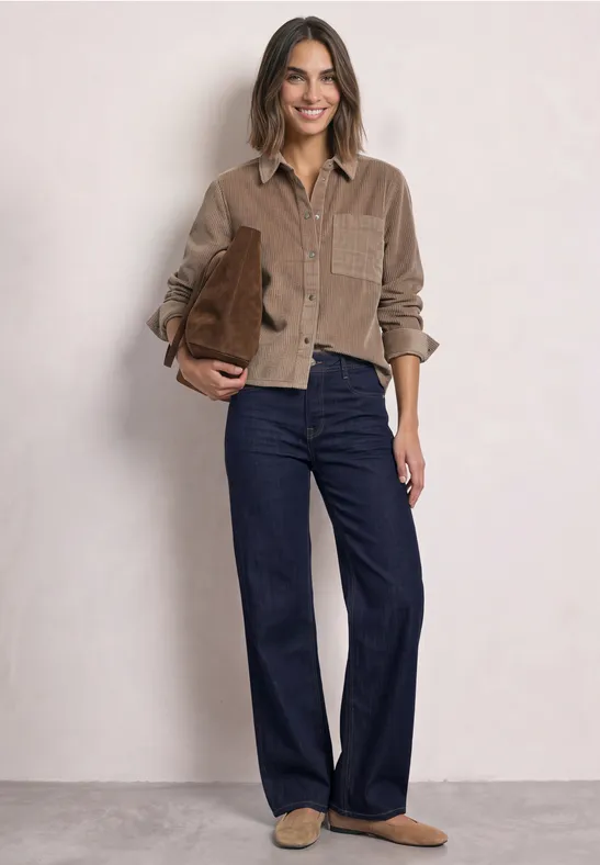 Eine Frau trägt ein braunes Cordhemd, weite Jeans und Hausschuhe, dazu eine braune Tasche.