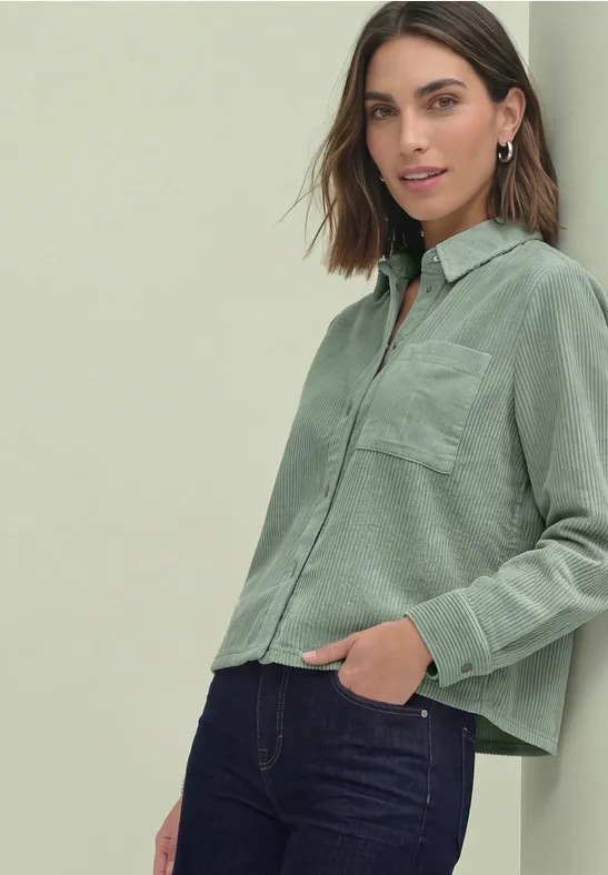 Frau trägt hellgrüne, locker sitzende Bluse mit Brusttasche und dunkle Jeans.