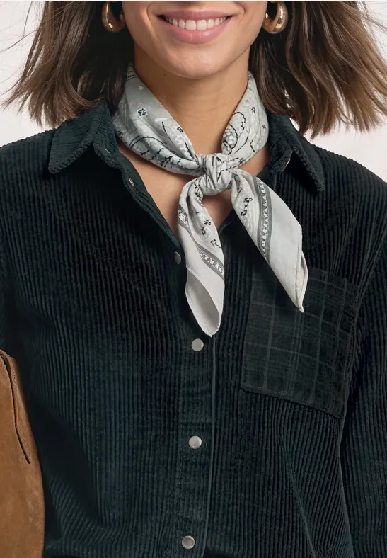 Vrouw draagt zwarte corduroy blouse en grijze sjaal om haar hals, met gouden oorbellen, glimlachend.
