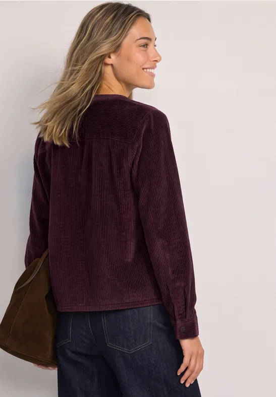 Frau von hinten mit violetter Cordjacke, blonden Haaren und Jeans, trägt eine Tasche über der Schulter.