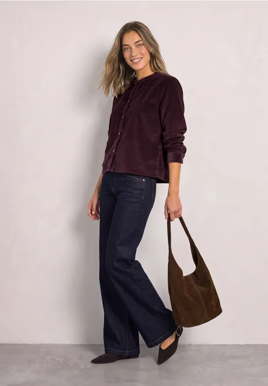 Eine Frau trägt eine dunkelviolette Samtjacke, breite Jeans, schwarze Schuhe und hält eine braune Wildledertasche.