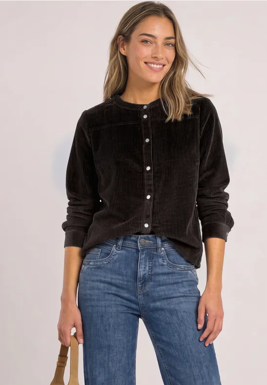 Junge Frau in schwarzem Samtblazer und blauer Jeans, lächelt und hält eine Tasche.